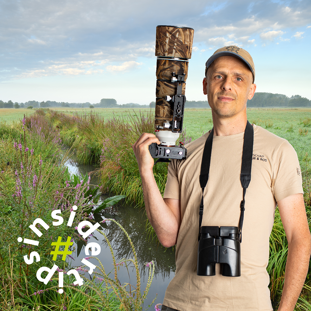 Dossierbehandelaar Kurt met groot natuurfototoestel in zijn hand en verrekijker rond zijn nek, en de Kalkense Meersen op de achtergrond